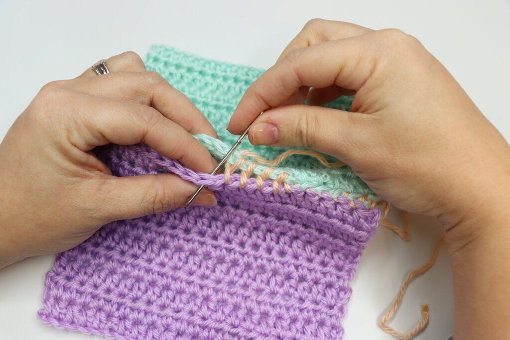 A pair of female hands is shown demonstrating how to to the mattress stitch. The swatch is purple, aqua and peach, bright colours so that the stitching is clearly demonstrated.