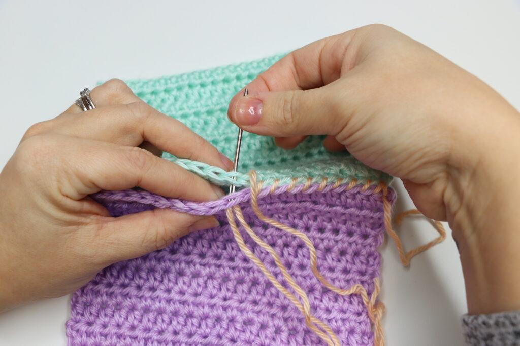 A pair of female hands is shown demonstrating how to to the whipstitch. The swatch is purple, aqua and peach, bright colours so that the stitching is clearly demonstrated.