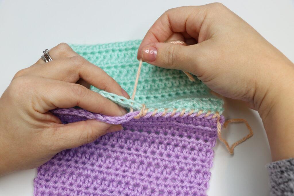 A pair of female hands is shown demonstrating how to to the whipstitch. The swatch is purple, aqua and peach, bright colours so that the stitching is clearly demonstrated.