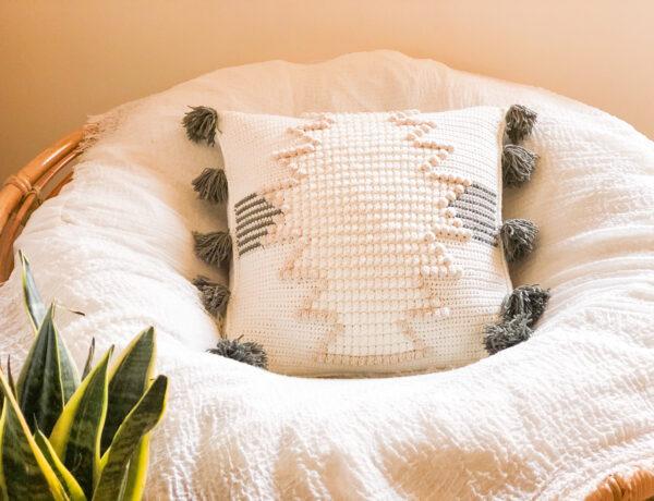 An aztec crochet cushion on a cosy chair in a living space next to a plant.