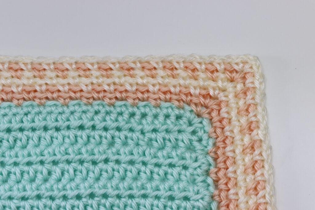A swatch of crochet sits on a white background. The moss stitch border is displayed around a plain crochet swatch in aqua yarn. The border is striped in peach and cream. 