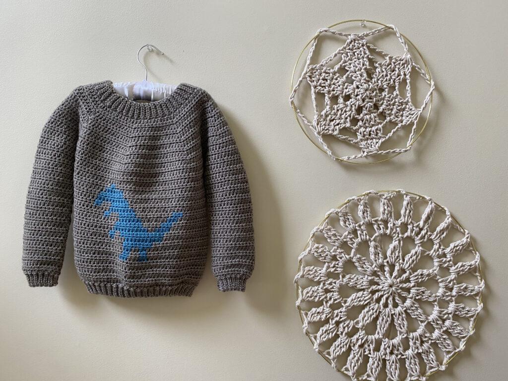 A crochet sweater made of brown yarn hangs on a white wall. The sweater has a blue dinosaur motif on the front. To the right is a pair of crochet mandalas in cream yarn.