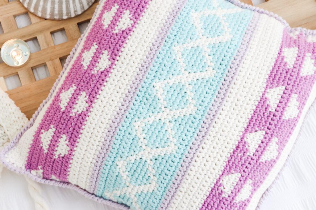 The Summer Breeze Tapestry Crochet Cushion is displayed on a white bed, propped against a wooden board beside shells and tealight candles. 