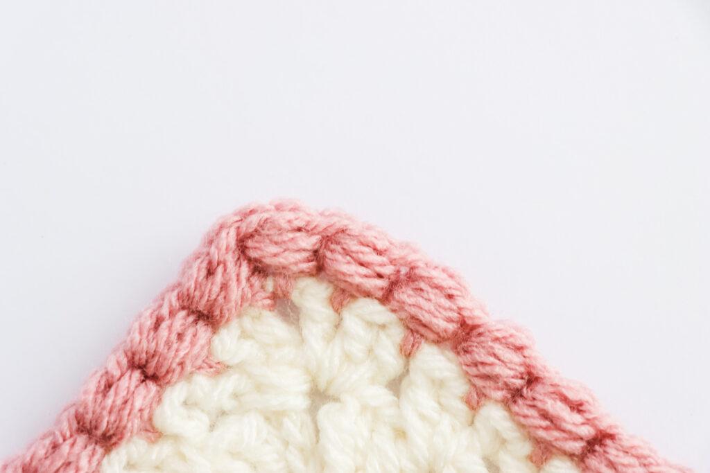 A swatch of puff edge border is displayed. The swatch is made from white yarn with the border made from a dusty rose colour. 