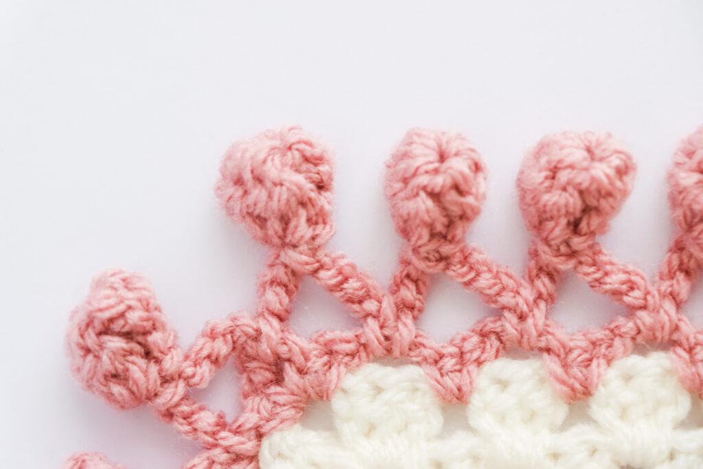 A swatch of pom pom border is displayed. The swatch is made from white yarn with the border made from a dusty rose colour. 