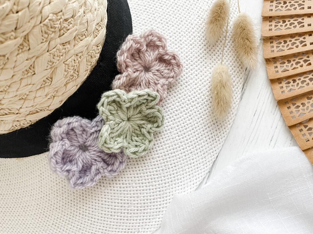 A trio of simple crochet flowers sit on the brim of a straw hat besides an open wooden fan.