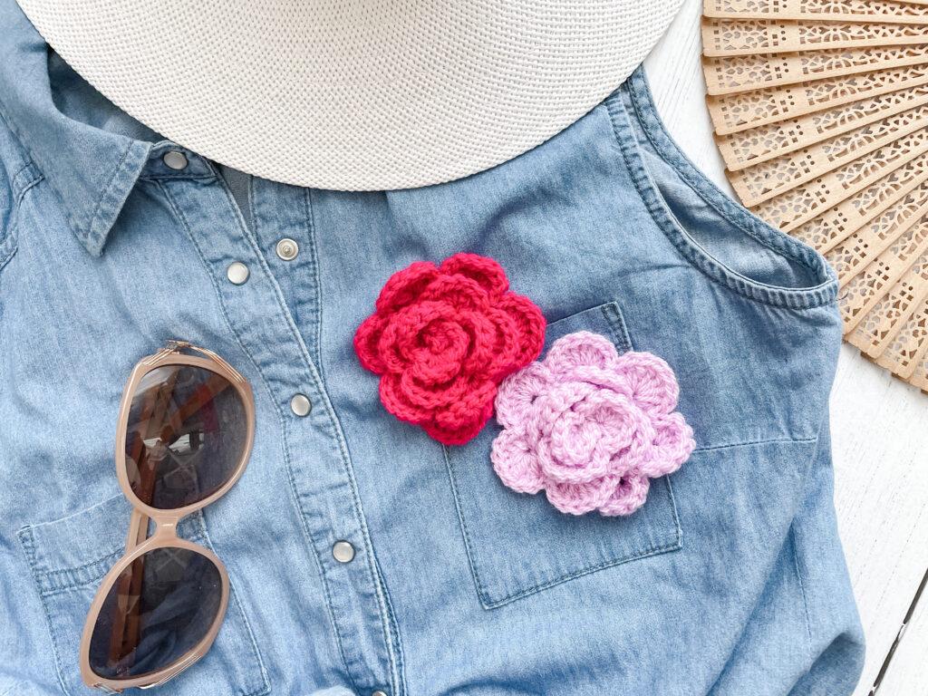 A pair of crochet roses in pale purple and bright pink are pinned to a denim vest in classic blue. A pair of brown sunglasses also sits on the vest, which is laid of a white wooden floor besides an opened wooden fan and a white hat brim.