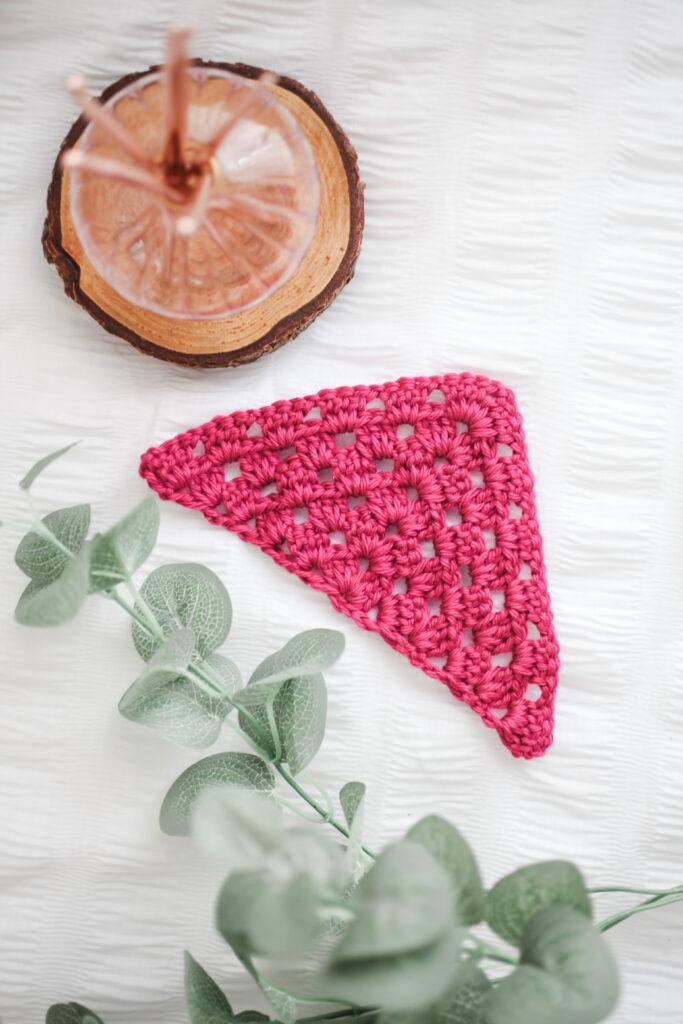 A triangular swatch of granny stitch in bright raspberry coloured yarn sits on a white background. Beside the swatch is a sprig of greenery. Above that, a slide of wooden log with a glass reed diffuser filled with reeds.