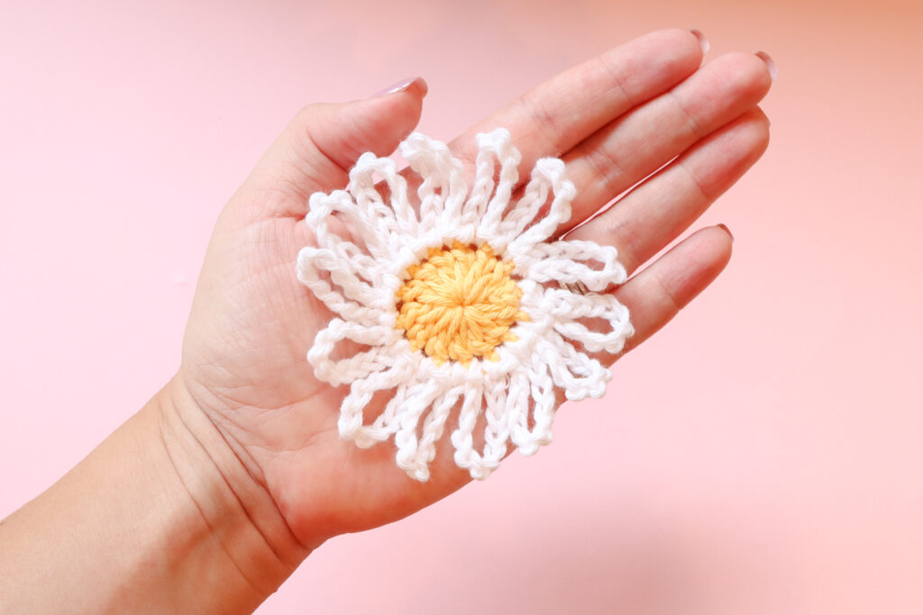 A white woman's hand holds a crochet daisy with a yellow centre and white petals. The background is pink.