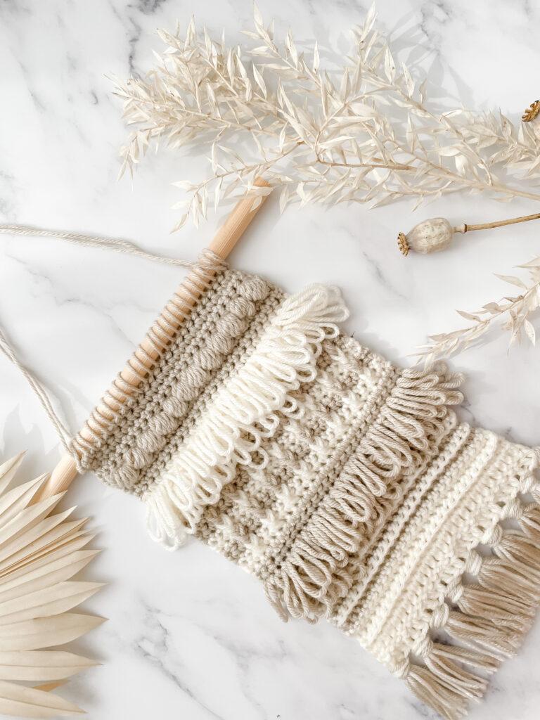 A crochet wall hanging made in cream and light brown yarn sits on a white marble background. The wall hanging is made up of different stitches. Shown here prominently are loop stitch, puff stitch and bar stitch. 