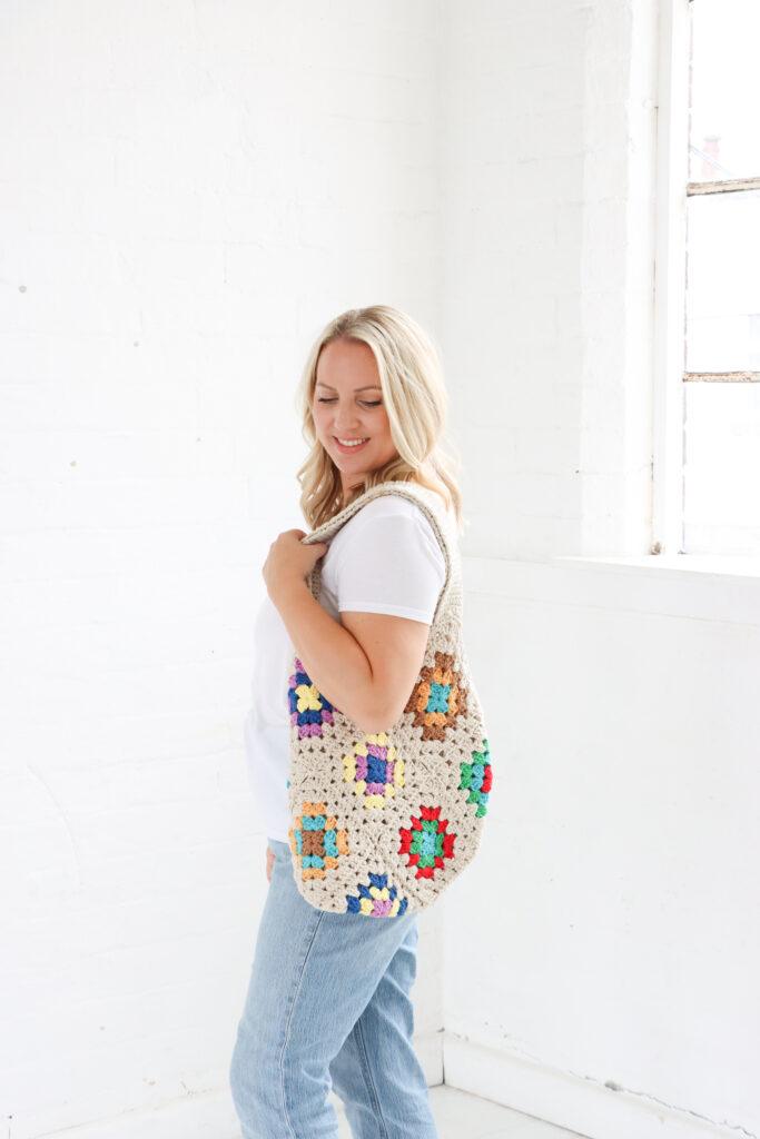 female stood in a white room wearing a white t-shirt and pale  blue jeans with a crochet granny square tote bag on her shoulder. 