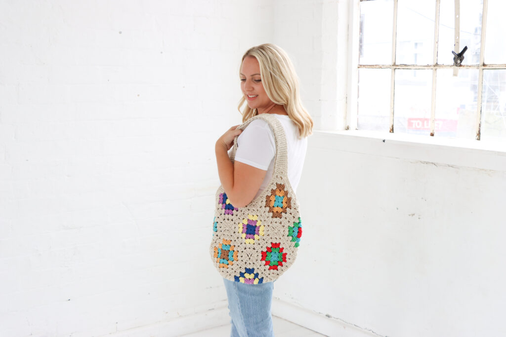 female wearing a white t-shirt and blue jeans in a white room with a granny square tote bag over her shoulder. 
