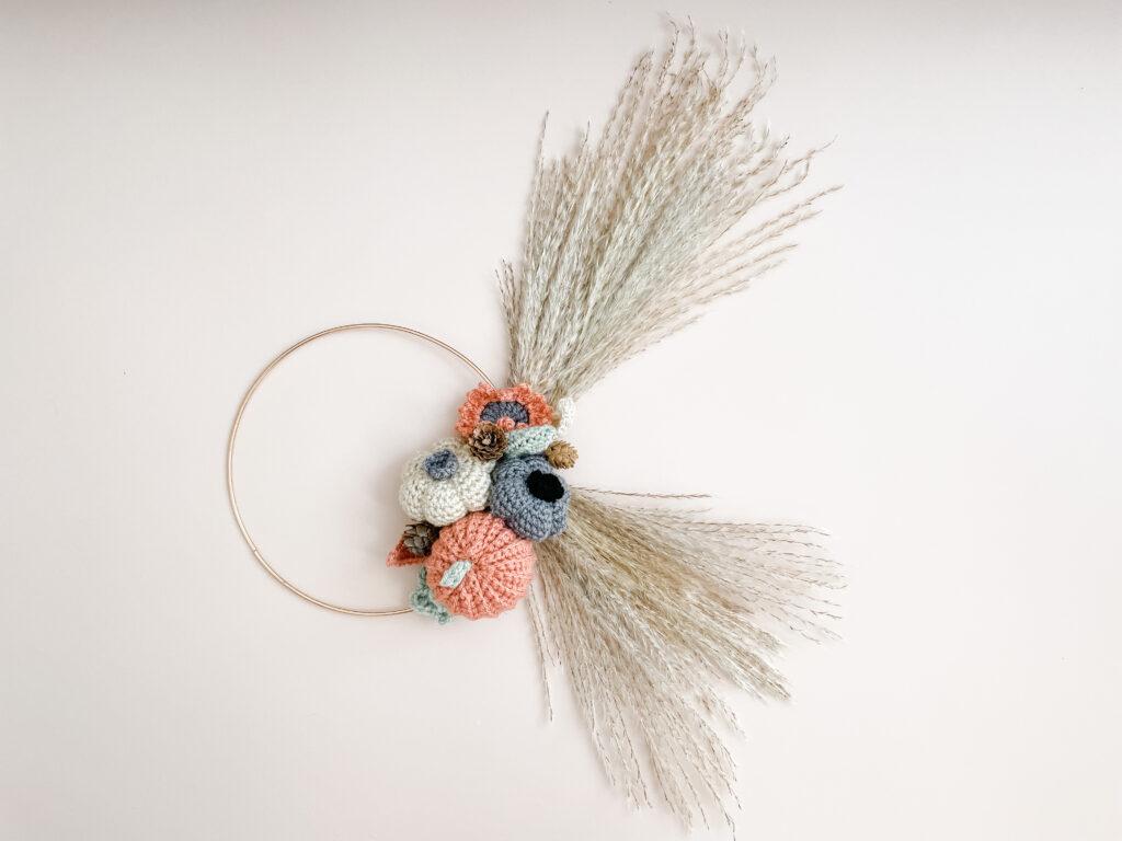 an flat lay image of a crochet wreath made of crochet flowers, pumpkin and leafs with pampas grass attached to a gold metal hoop on a pale pink back ground. 