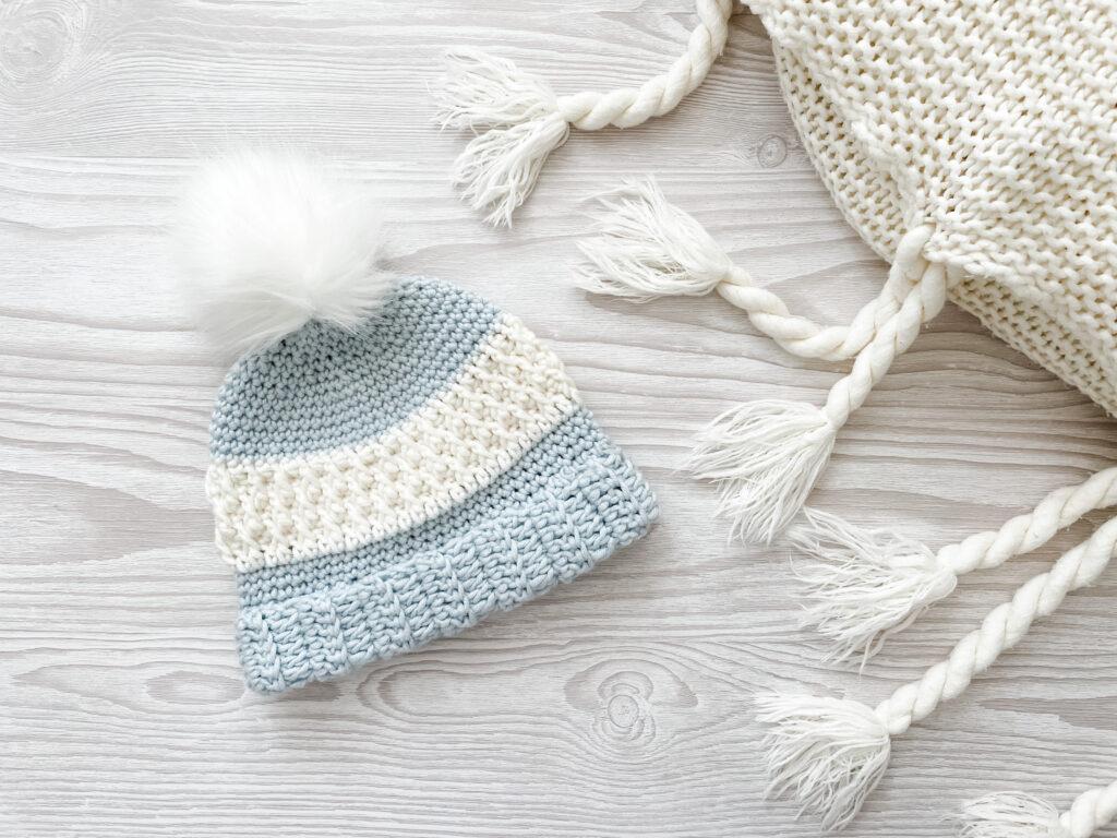 The Alaskan Delights hat sits on  a pale grey wooden background. Made from a pale blue and white yarn combination and topped with a white fluffy pompom, the hat is most prominently made using the alpine stitch.