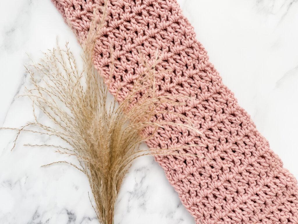 The Easy Infinity Scarf is made in dusty pink yarn. It sits draped over a white marble background beside some pampas grass.