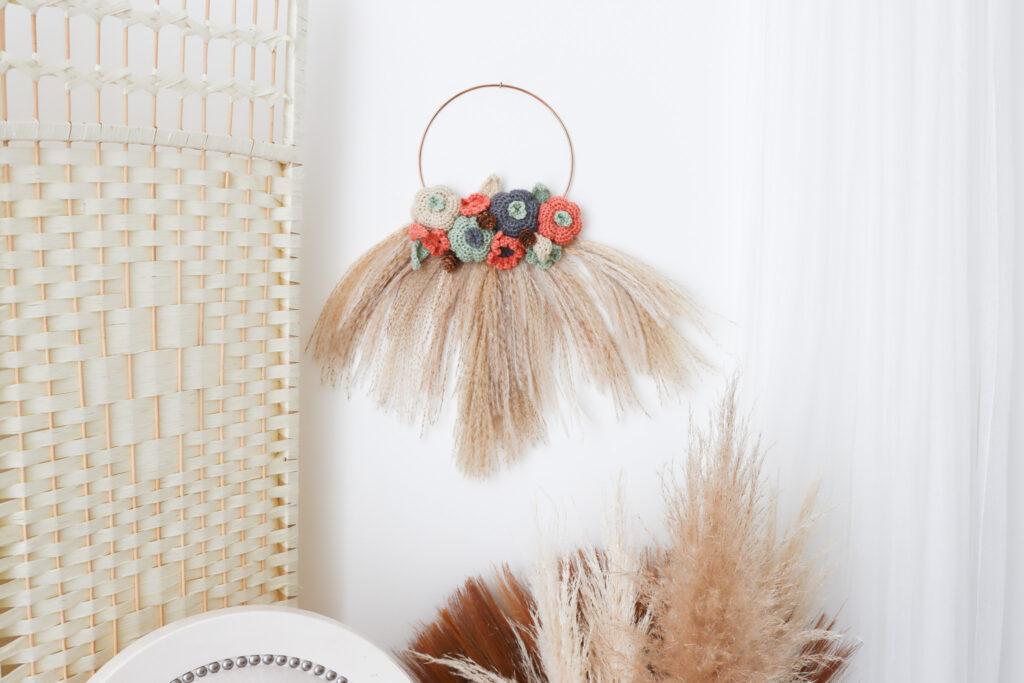 an image of a crochet wreath made of crochet flowers, pumpkin and leafs with pampas grass hung on a white wall with a wicker room divider to the left. There is a white chair underneath the wreath and more pampas grass to the right with a white net curtain. 