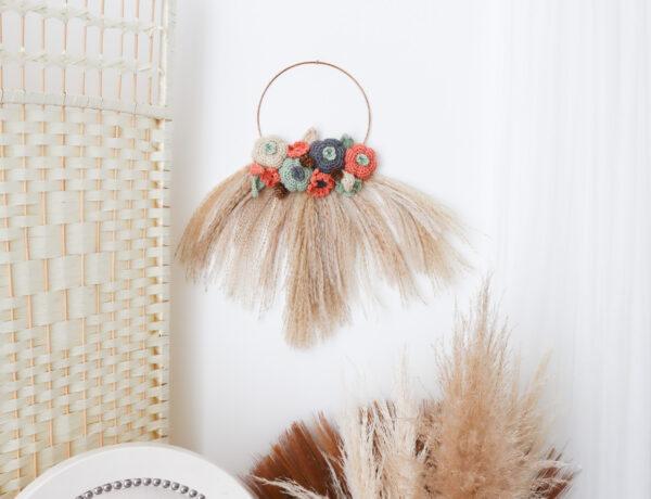 an image of a crochet wreath made of crochet flowers, pumpkin and leafs with pampas grass hung on a white wall with a wicker room divider to the left. There is a white chair underneath the wreath and more pampas grass to the right with a white net curtain.