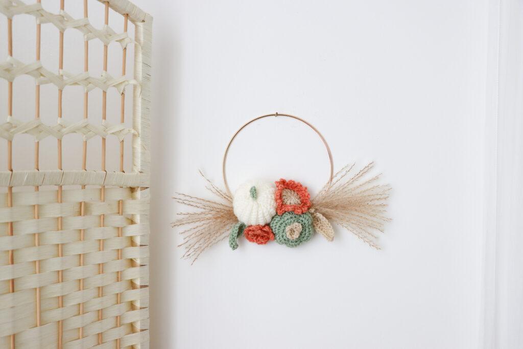 an image of a crochet wreath made of crochet flowers, pumpkin and leafs with pampas grass hung on a white wall with a wicker room divider to the left. 