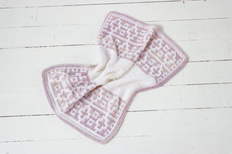 crochet mosaic baby blanket in soft pastel shades of white, pink, and lilac, laid flat on a white wooden floor