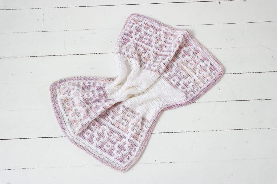 crochet mosaic baby blanket in soft pastel shades of white, pink, and lilac, laid flat on a white wooden floor