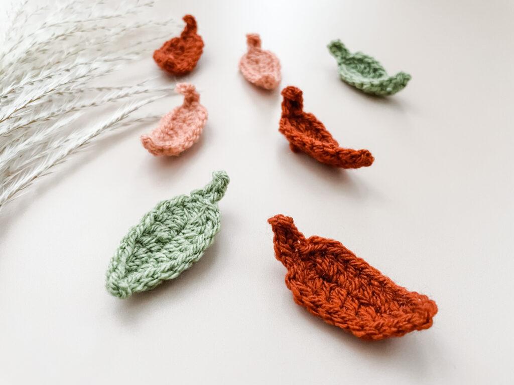 a flat lay image of 7 crochet leaves in green, rust and peach yarn lay on a cream backdrop with pampas grass appearing on the left of the image. 