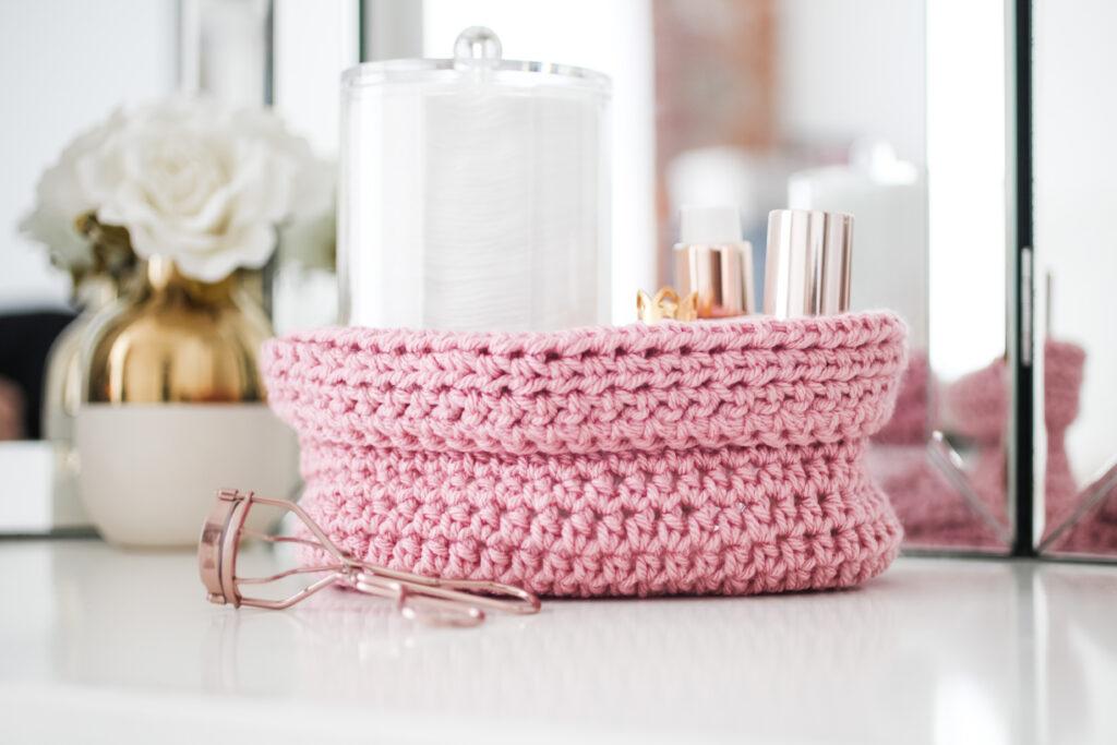 A crochet oval basket with make up and creams in on a dressing table next to an eyelash curler.