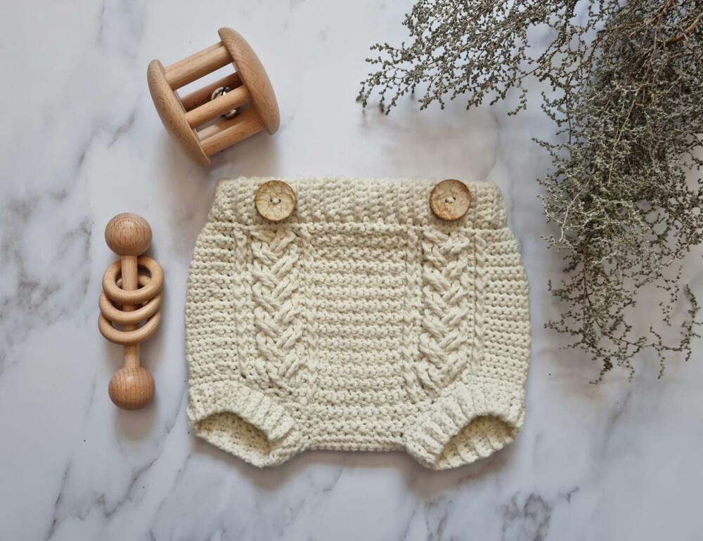 crochet baby bloomers lay flay on a marble backdrop with two wooden toys and foliage in the top right corner of the image