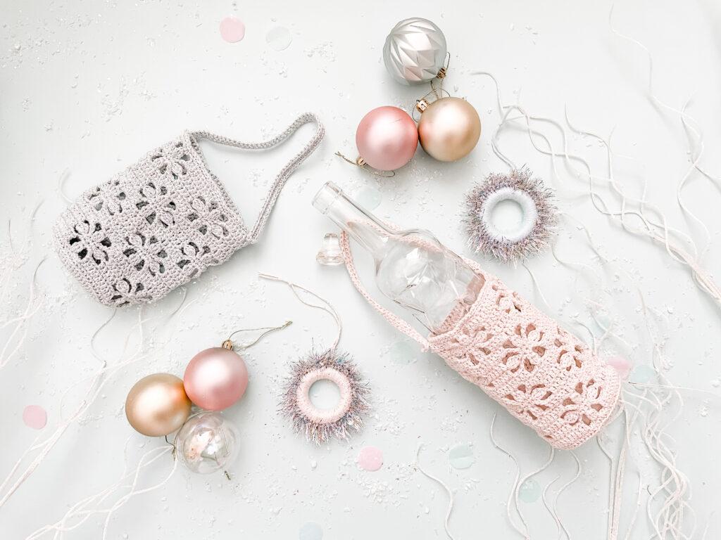 a flat lay image of two crochet bottle wrpas made in cream and silver sparkly yarn, one with an empty glass bottle in to show how it looks. lay around the bottle wraps are baubles, two present toppers and sparkly silver branches.