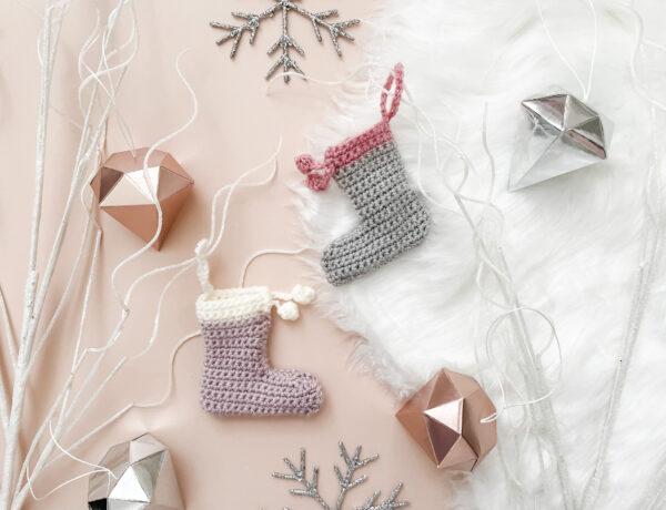 two mini crochet stockings lay flat on a pale pink backdrop with a fur rug to the right hand side of the image surrounded by white sparkly branches, snowflake and diamond-shaped decorations