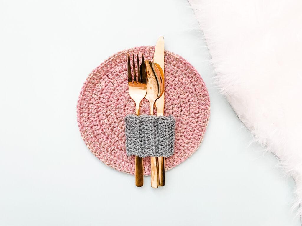 a crochet placemat made in pink and gold yarn on a pale blue backdrop. Lay on the placemat is some gold cutlery wrapped in a silver crochet cutlery holder/wrap.