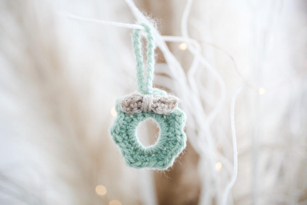 crochet mini wreath hung from a white branch with fairy lights in the background. the mini wreath is made in pale green yarn with a cream bow. 
