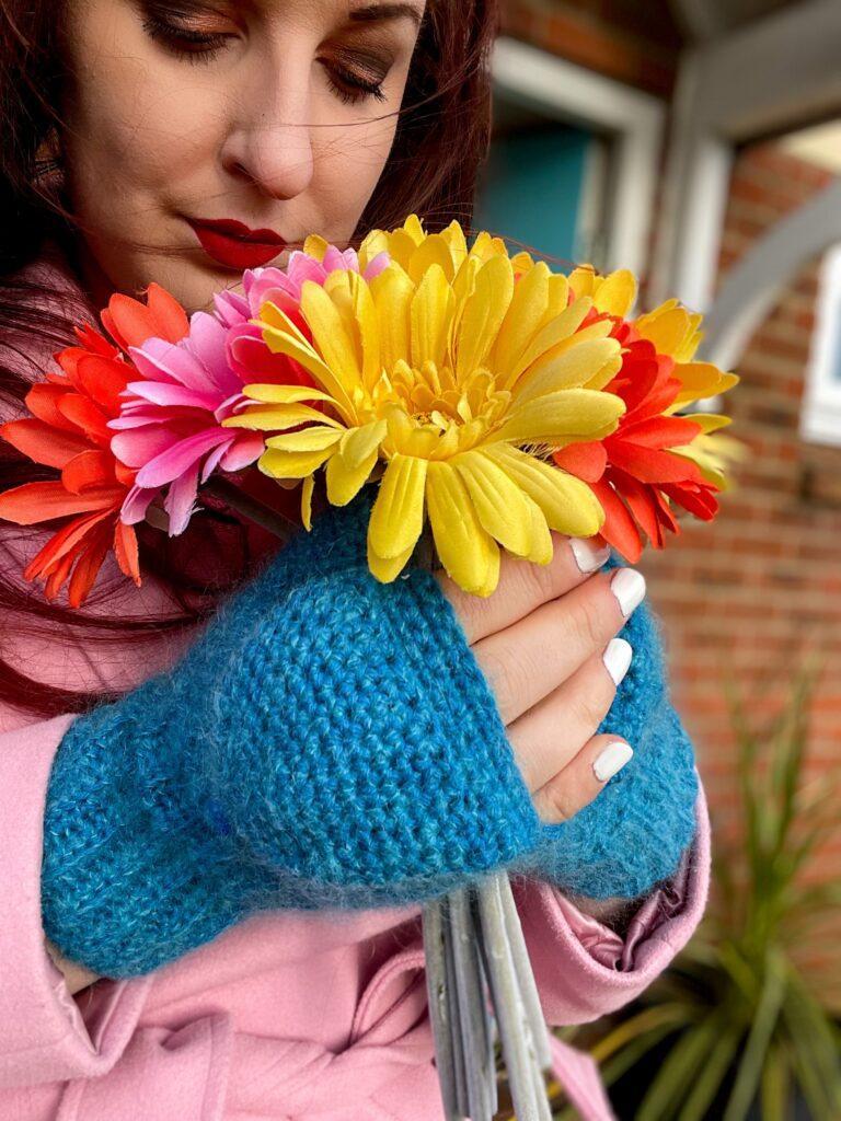 a woman holding a bunch of bright yellow and orange flowers wearing blue crochet mittens and a pink coat. 