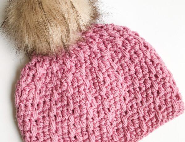 a pink crochet beanie hat with a camel coloured fur pom pom lay flat on a white background