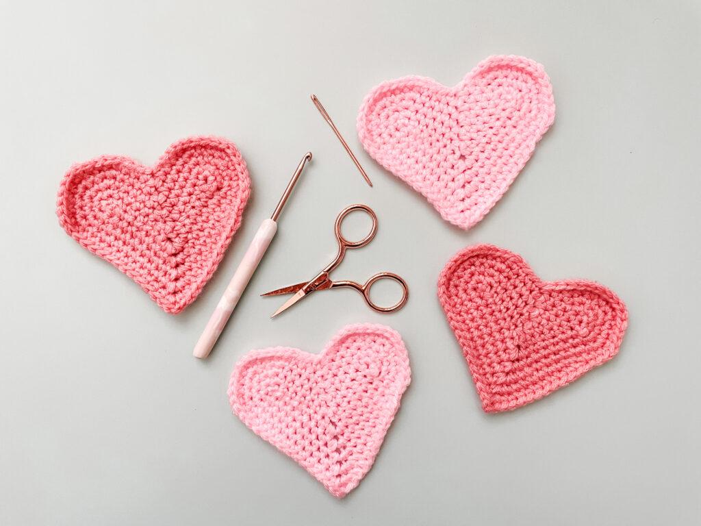 flat lay image of four heart coasters on a pale blue background  with a rose gold crochet hook, scissors and darning needle.
