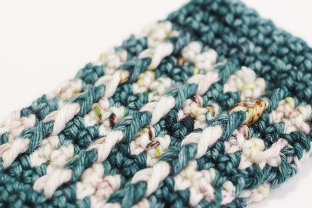 a close-up image of a crocheted wrist warmer in green and cream indie-dyed yarn. The image shows the texture of the bar stitch using indie-dyed yarn. 