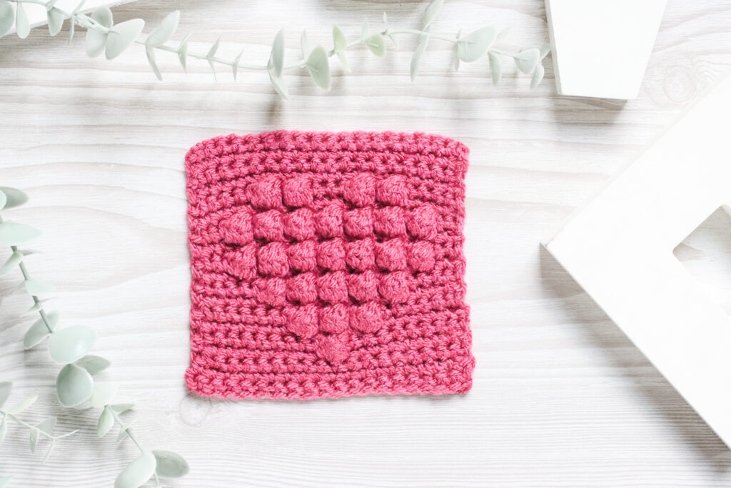 A fuchsia granny square in pink yarn on a wooden table with plant vine surrounding it.