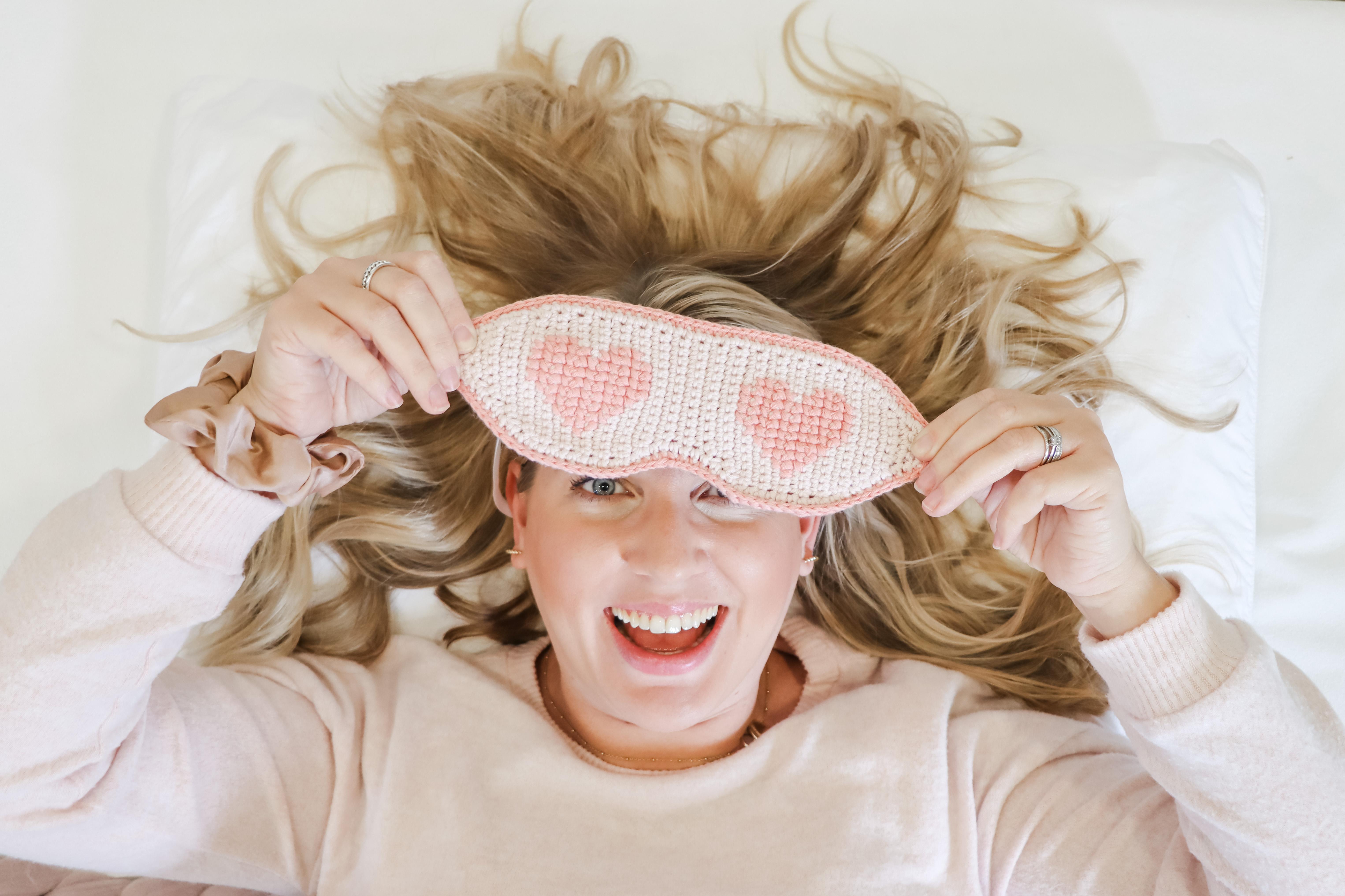 Female lay down holding a crochet sleep mask out in front of her face covering one eye. 