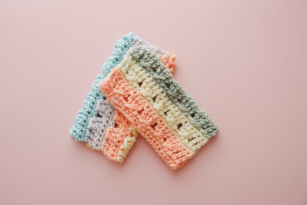 a pair of crochet wrist warmers lay crossed over each other on a pink backdrop.v