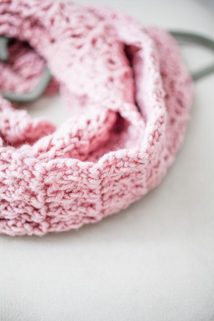 a close-up image of a pink crochet infinity scarf wrapped around a coast hanger lay flat on a cream chair. 
