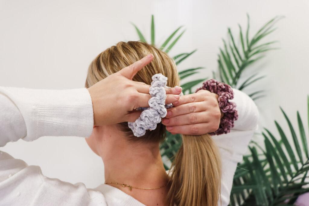 A girl putting her hair up into a ponytail with a handmade crochet scrunchie made from grey velvet yarn.