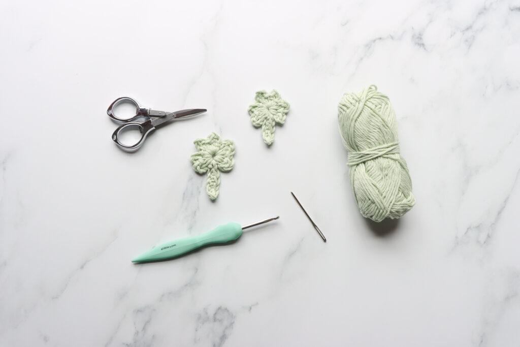 a flat lay of equipment with what you will need to make a crochet shamrock. Including in the picture is a ball of pale green yarn, two crochet shamrocks, scissors, crochet hook and darning needle. 