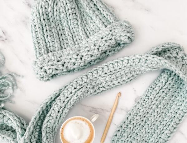 aA crochet hat and scarf made in duck egg yarn lay flat on a marble backdrop. There is a coffee and a bamboo crochet hook is lay in between the scarf.