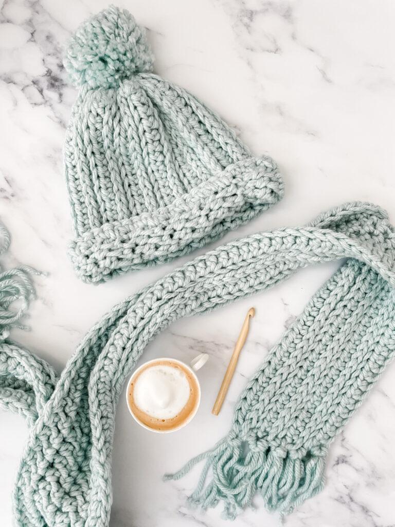 aA crochet hat and scarf made in duck egg yarn lay flat on a marble backdrop. There is a coffee and a bamboo crochet hook is lay in between the scarf. 