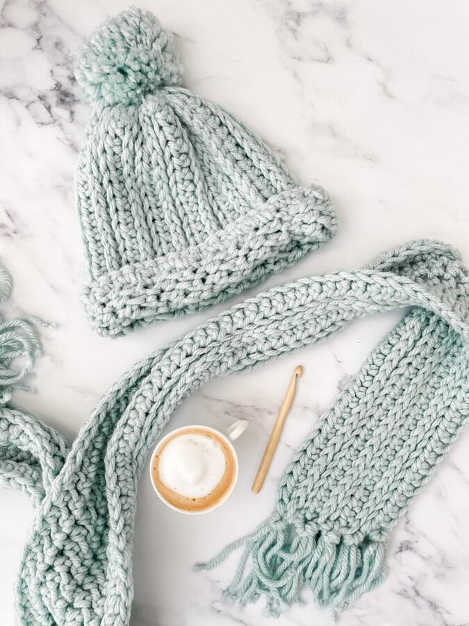 aA crochet hat and scarf made in duck egg yarn lay flat on a marble backdrop. There is a coffee and a bamboo crochet hook is lay in between the scarf.