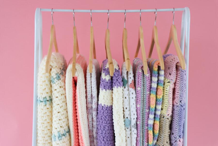 Colourful handmade crochet blankets hanging neatly on wooden hangers against a pink background.