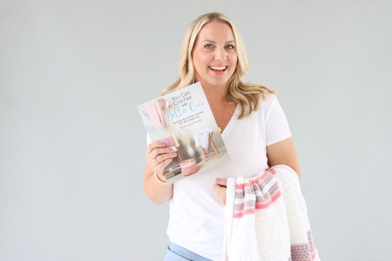 Sarah-Jayne from Bella Coco stood infont of a grey background with a crochet blanket draped over her arm whilst holding her new book in the opposite hand.