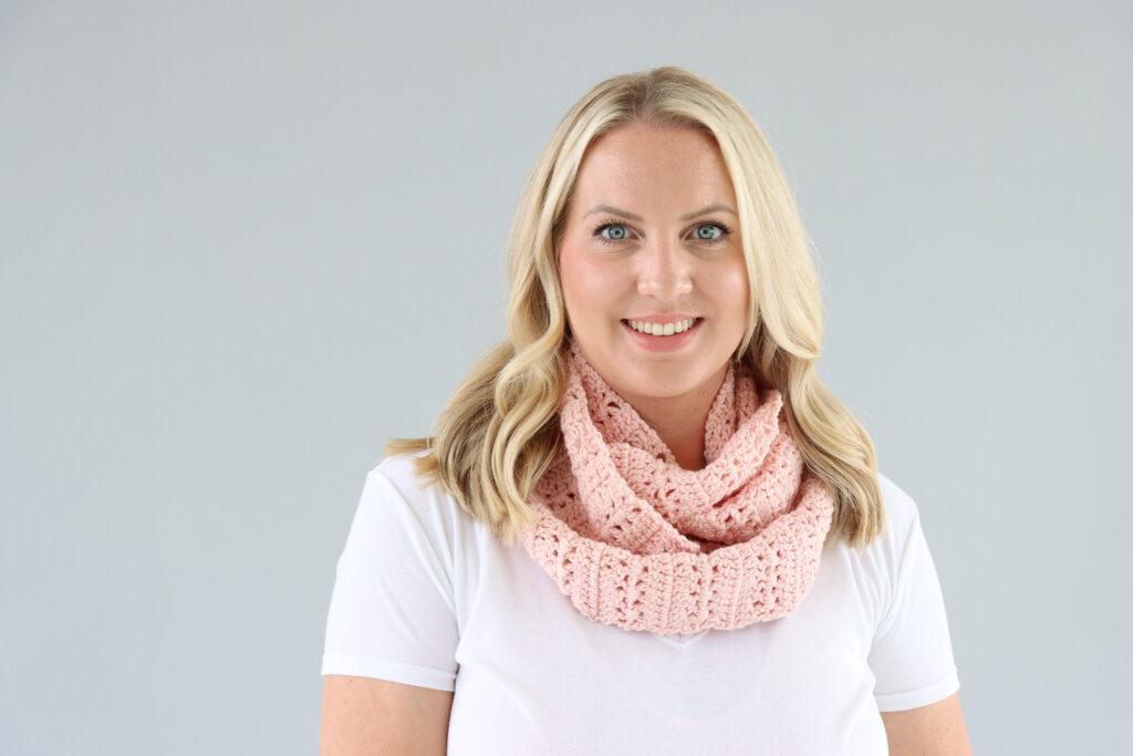 female stood in front of a grey background wearing a white t.shirt and a pale pink crochet infinity scarf. 