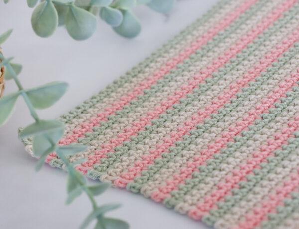 a swatch of UK double crochet using three colours of yarn in paintbox cotton dk in colours: pistachio, vanilla cream and blush pink. The swatch of double crochet is lay on a marble background with a green trailing palm tin the top left corner of the image.