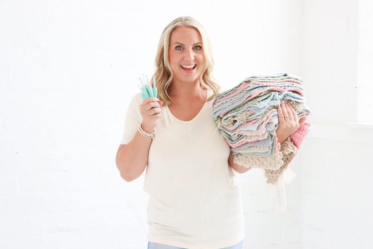 A happy woman holding a stack of crochet squares for a patchwork blanket in her arms. She's holding a crochet hook in the other hand.