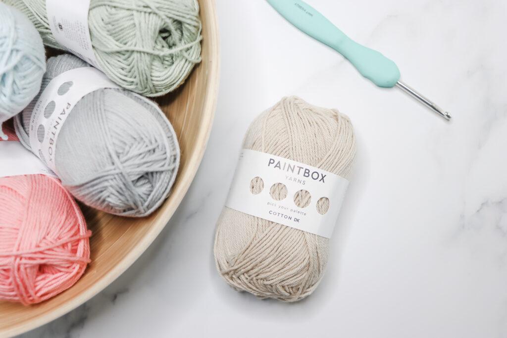 An image featuring a bowl of pastel colours yarn set to the left of the image. To the right of the image is a ball of cream yarn with an Athena crochet hook above the ball of yarn lay on a marble background.